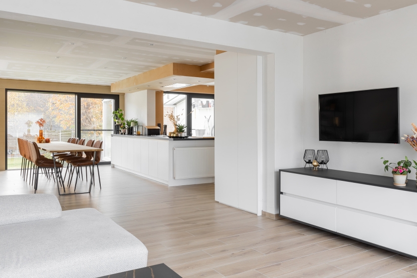 Moderne, open woonkamer en keuken met houten vloer, strakke witte kasten en grote ramen in een gerenoveerde woning