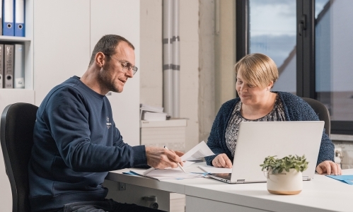Twee mensen bespreken renovatieplannen aan bureau met laptop en documenten in lichte kantoorruimte