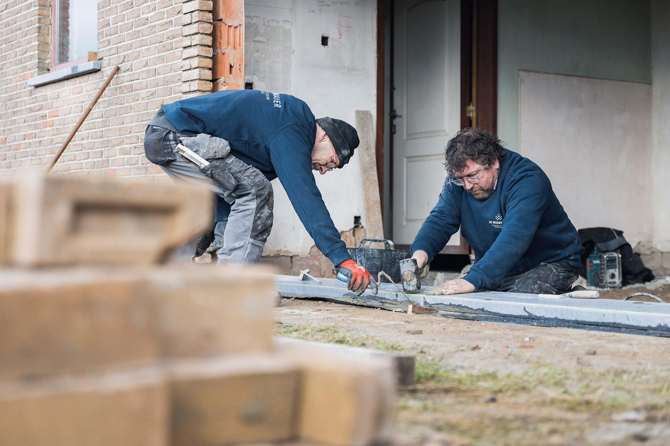 Twee vakmannen van De Woonwaaier bezig met renovatiewerken aan een woning, inclusief metselwerk en ruwbouw