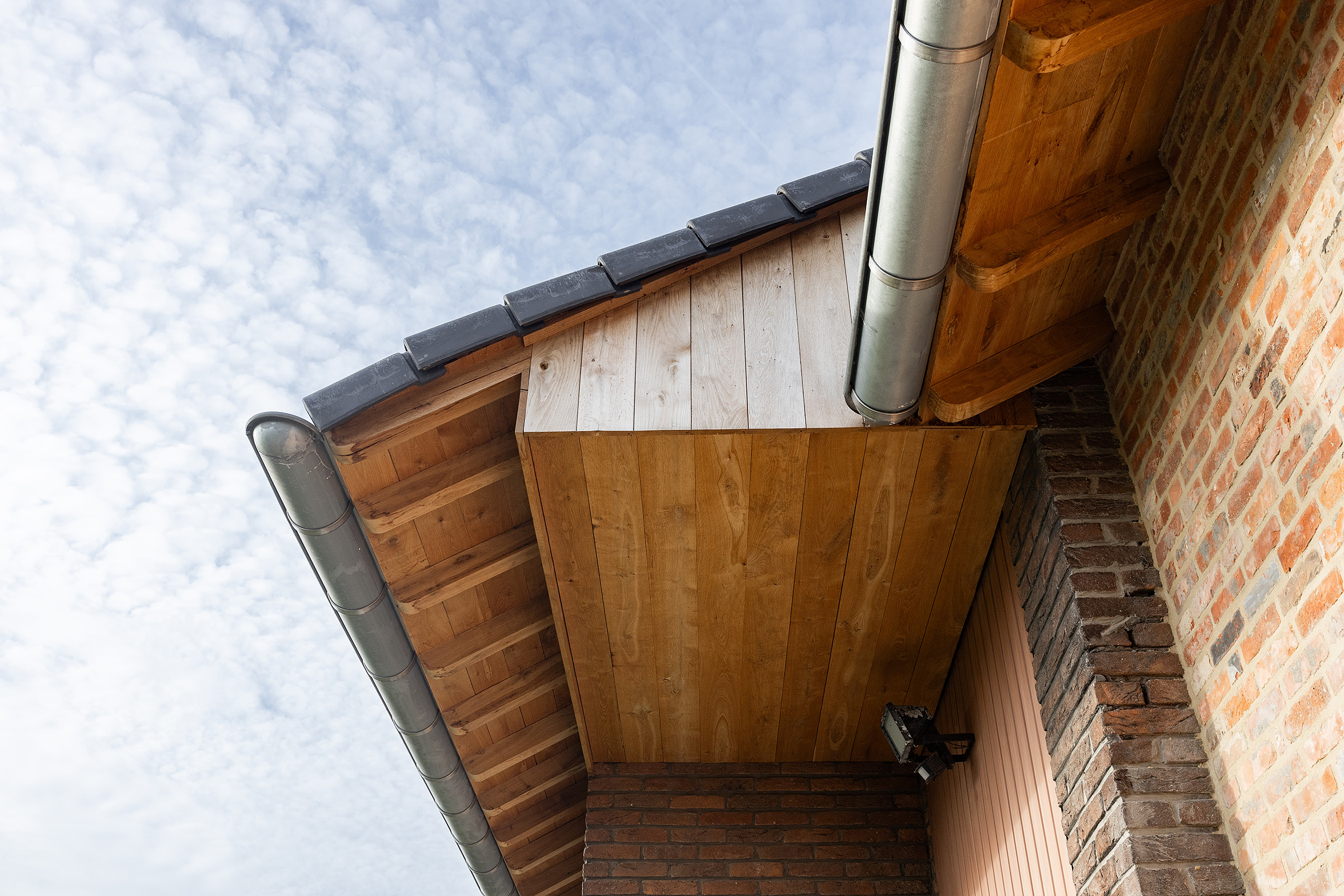 Dakoverstek met houten afwerking en metalen dakgoot aan bakstenen woning tegen een blauwe lucht met wolken