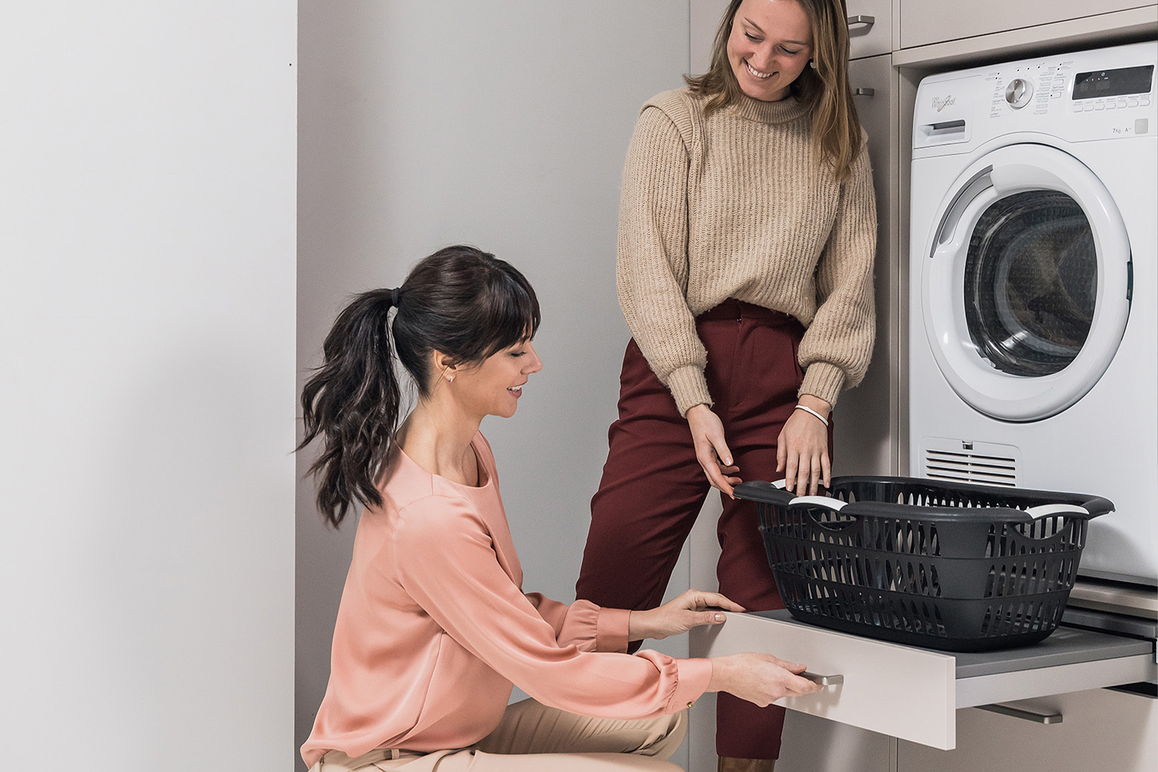 Twee vrouwen in een moderne wasruimte met wasmachine en kastje, bezig met huishoudelijke taken