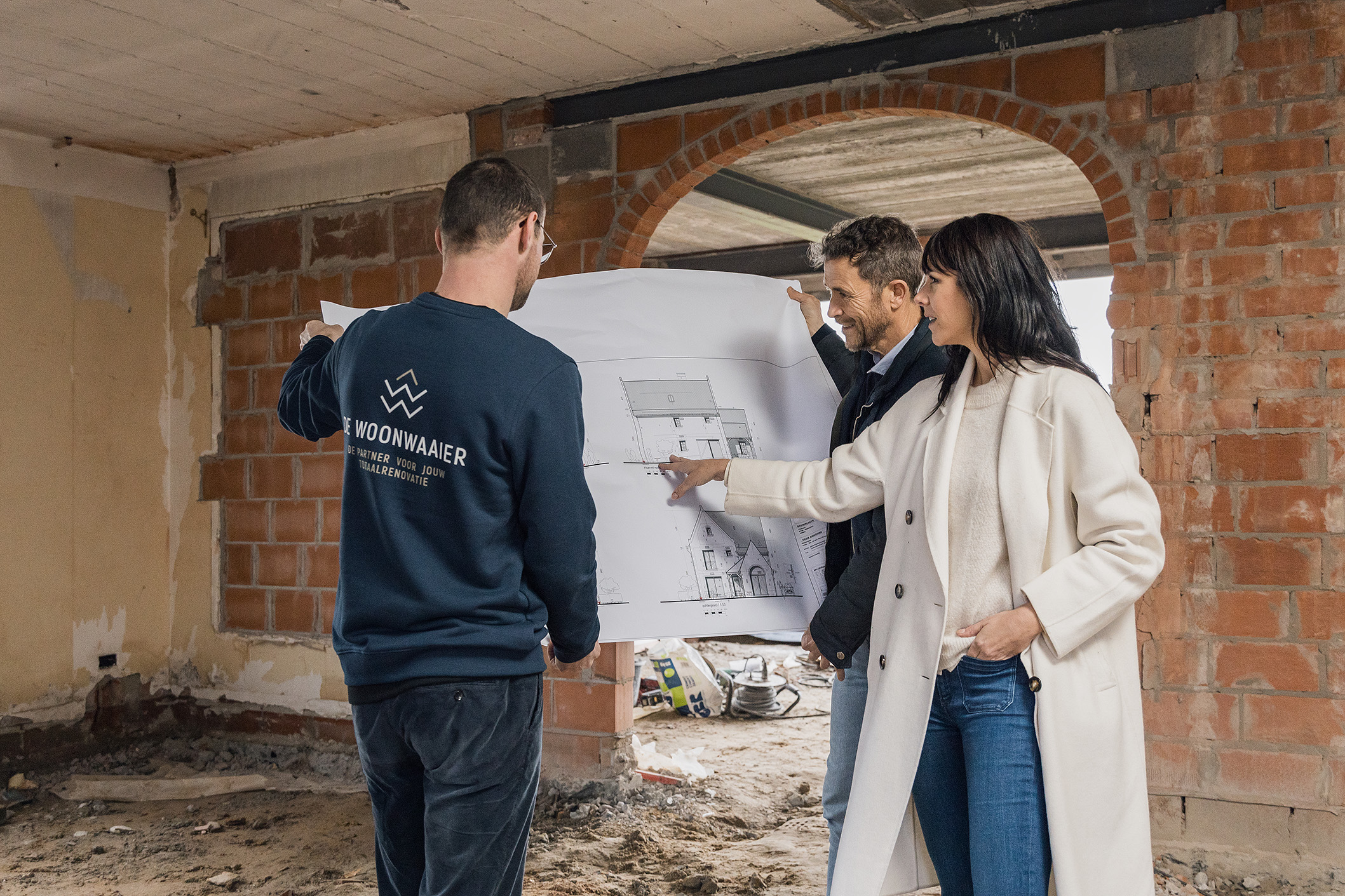 Drie personen bekijken bouwplannen in een woning in renovatie met zichtbare bakstenen muren en betonnen plafond