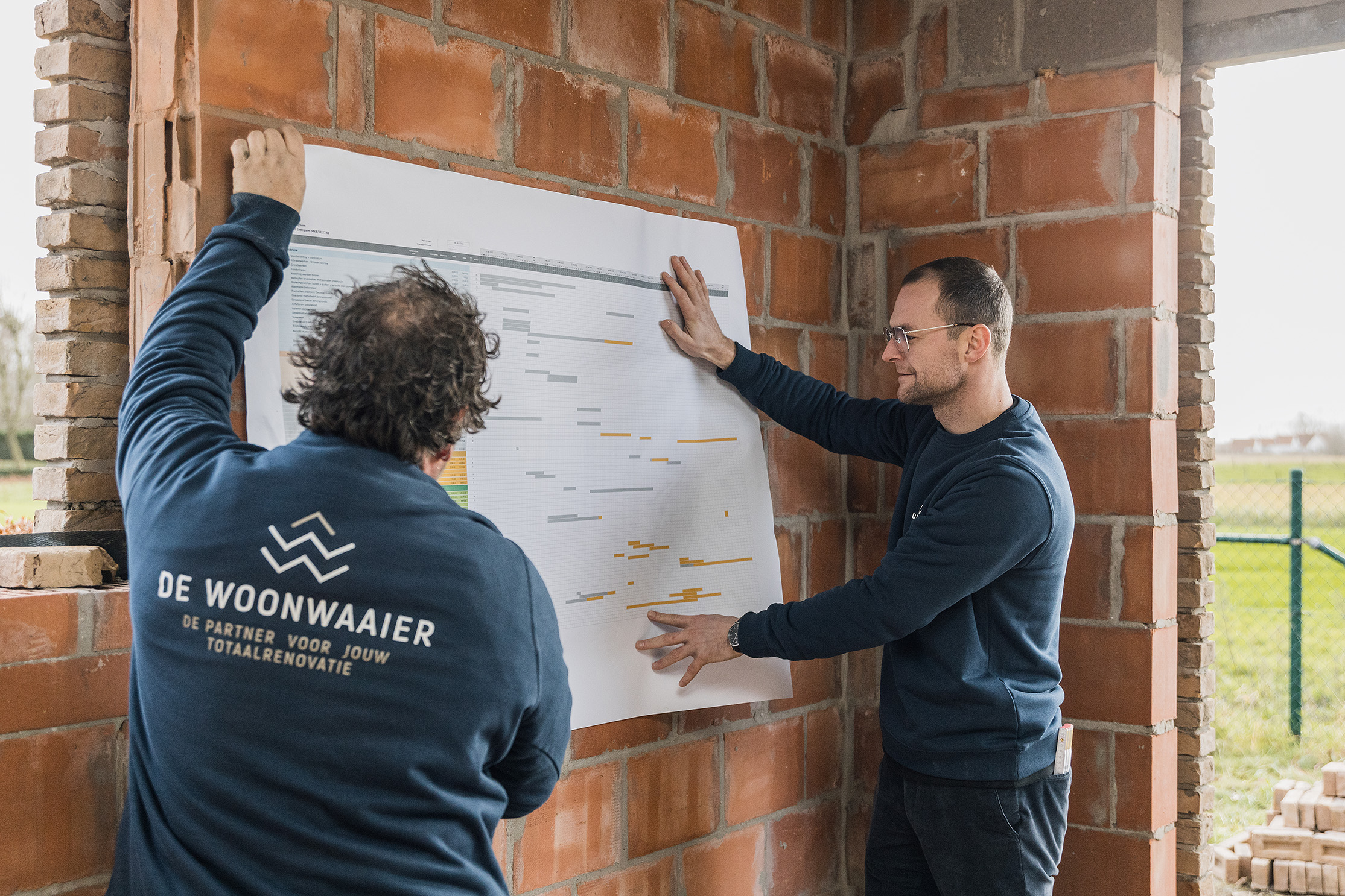 Twee mannen bespreken renovatieplannen van De Woonwaaier op een bouwplaats met zichtbare bakstenen muren